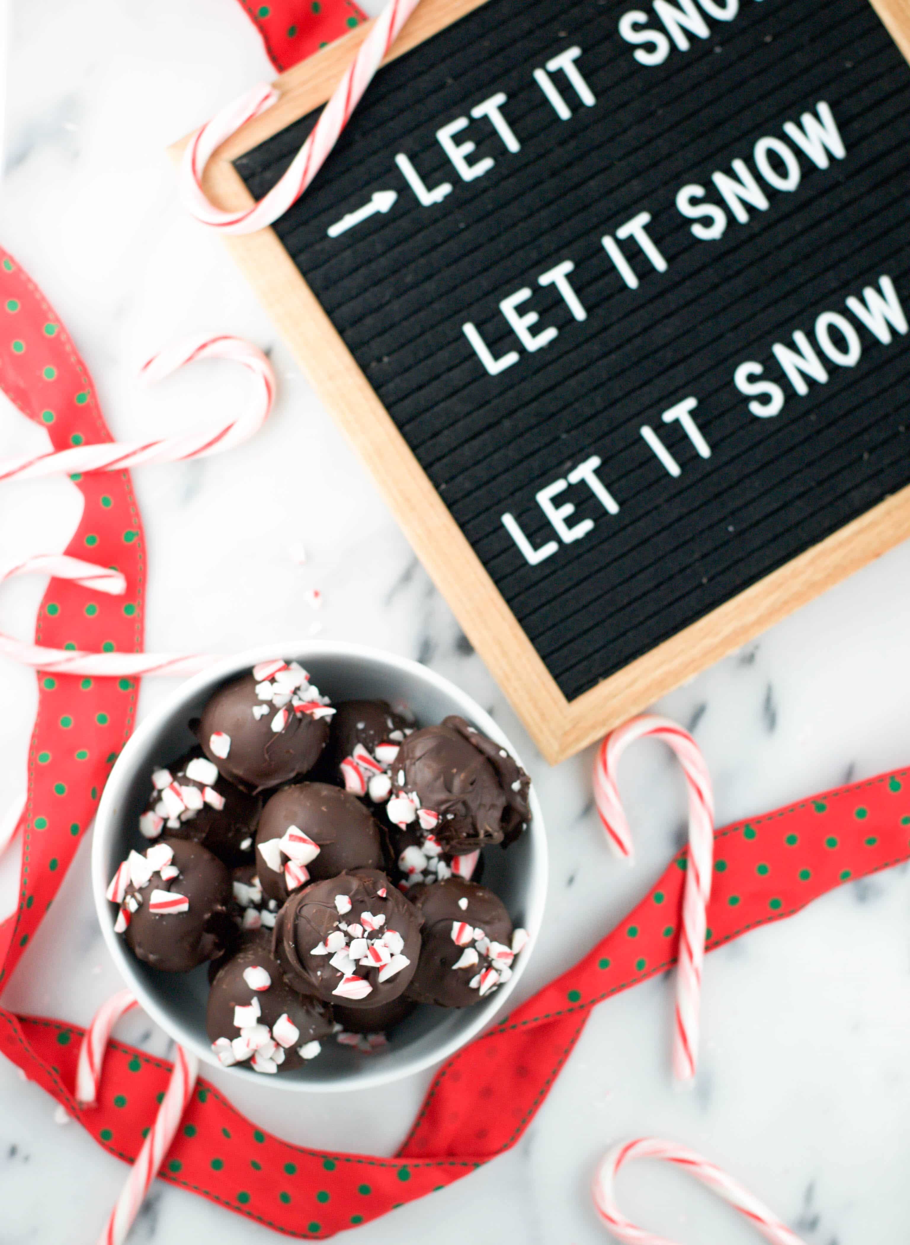 A bowl of peppermint truffles with a letterboard reading "Let it Snow, Let it Snow, Let it Snow" and some candy canes on the surface. 