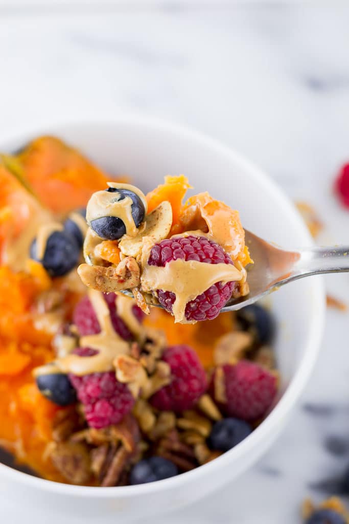 Baked Sweet Potato Breakfast Bowl with Berries - White bowl with baked sweet potato topped with creamy almond butter, fresh raspberries and blueberries and paleo granola including coconut, cashews, almonds, sunflower seeds, cinnamon and coconut oil