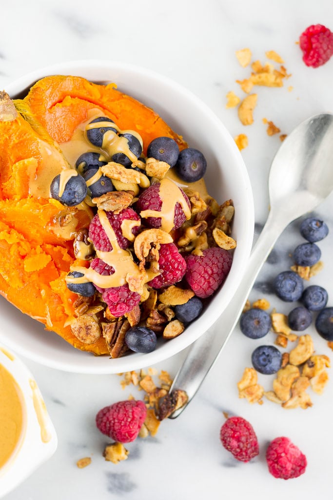 Baked Sweet Potato Breakfast Bowl with Berries - White bowl with baked sweet potato topped with creamy almond butter, fresh raspberries and blueberries and paleo granola including coconut, cashews, almonds, sunflower seeds, cinnamon and coconut oil