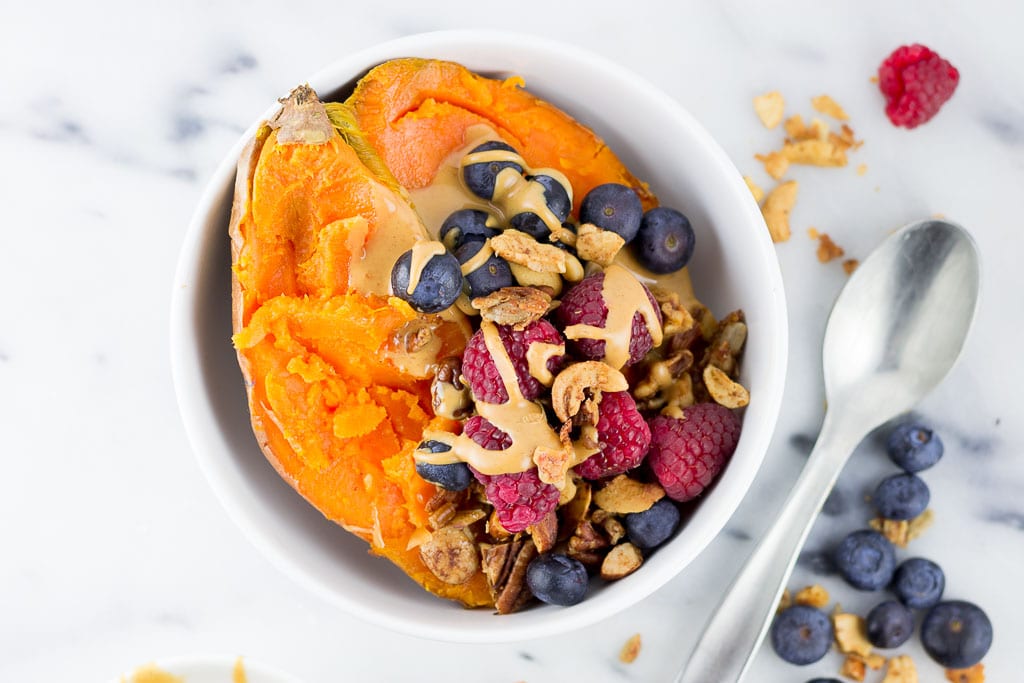 Baked Sweet Potato Breakfast Bowl with Berries - White bowl with baked sweet potato topped with creamy almond butter, fresh raspberries and blueberries and paleo granola including coconut, cashews, almonds, sunflower seeds, cinnamon and coconut oil