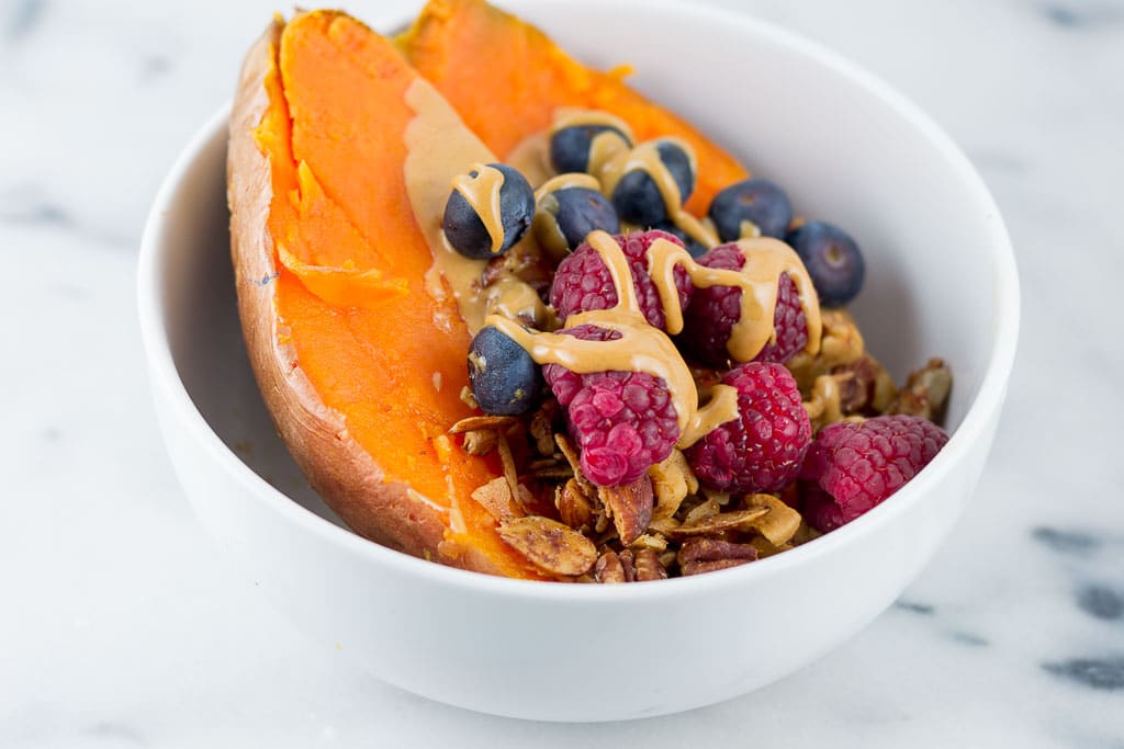 Baked Sweet Potato Breakfast Bowl with Berries - White bowl with baked sweet potato topped with creamy almond butter, fresh raspberries and blueberries and paleo granola including coconut, cashews, almonds, sunflower seeds, cinnamon and coconut oil