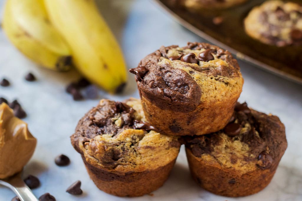 chocolate peanut butter banana muffins in a muffin tin with a spoonful of peanut butter and scattered dark chocolate chips on a marble table - gluten-free, vegan, dairy-free swirl muffin