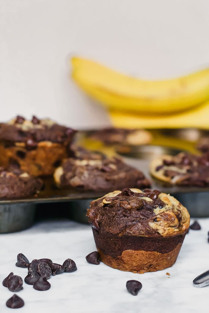 chocolate peanut butter banana muffins in a muffin tin with a spoonful of peanut butter, a bunch of bananas and scattered dark chocolate chips on a marble table - gluten-free, vegan, dairy-free swirl muffin