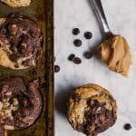 chocolate peanut butter banana muffins in a muffin tin with a spoonful of peanut butter and scattered dark chocolate chips on a marble table - gluten-free, vegan, dairy-free swirl muffin