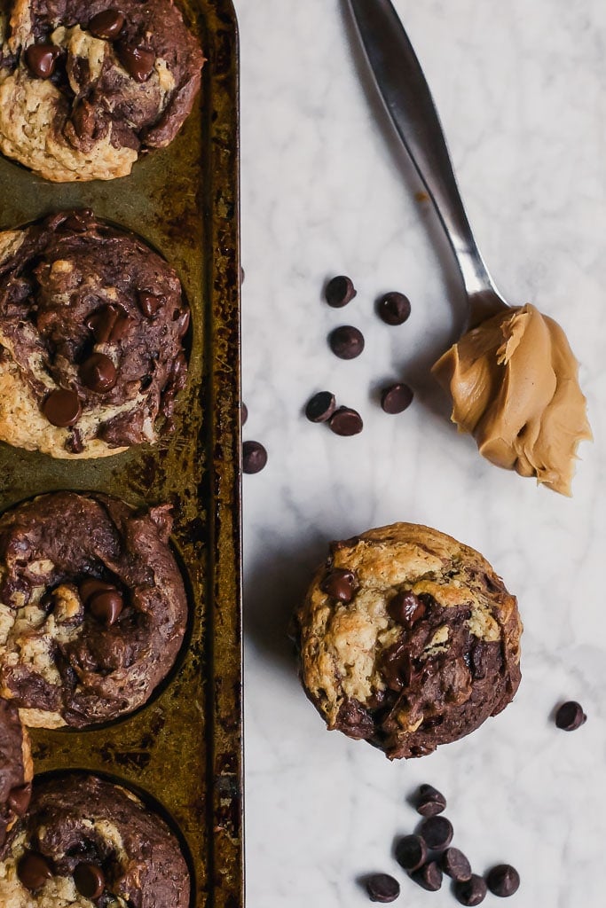 chocolate peanut butter banana muffins in a muffin tin with a spoonful of peanut butter and scattered dark chocolate chips on a marble table - gluten-free, vegan, dairy-free swirl muffin