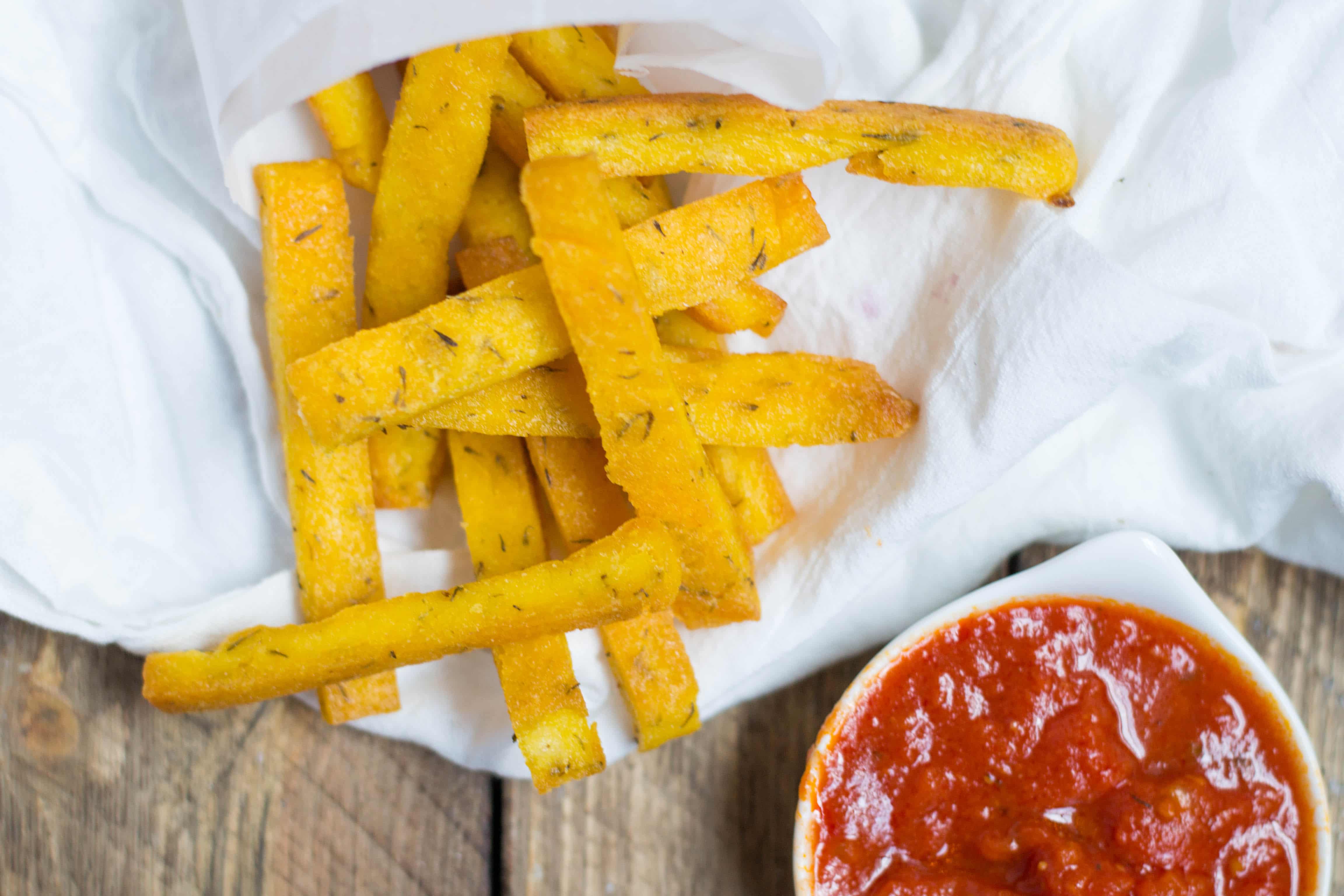 A parchment paper cone filled with polenta fries and a small bowl of marinara sauce. 