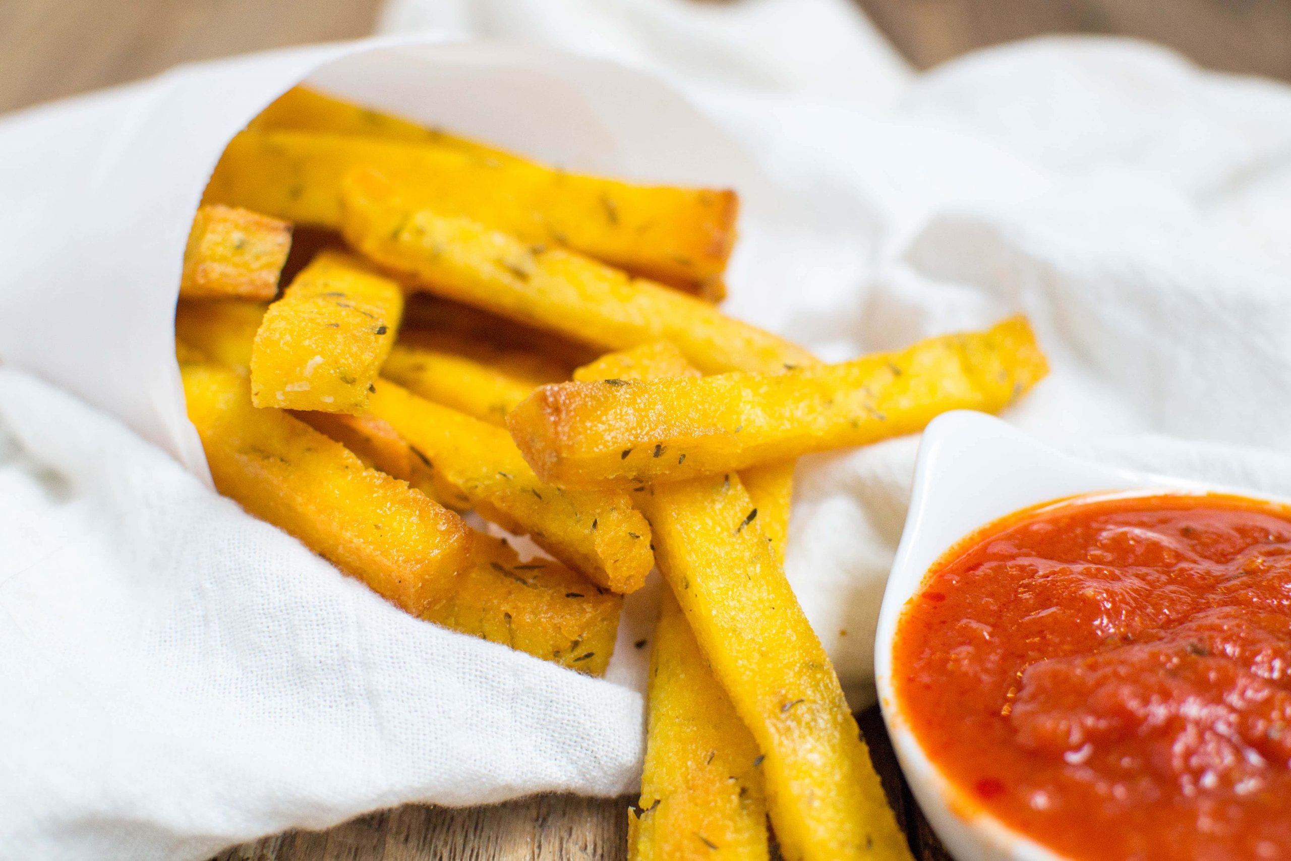 A cone of parchment paper with polenta fries coming out of it and a small bowl of marinara. 