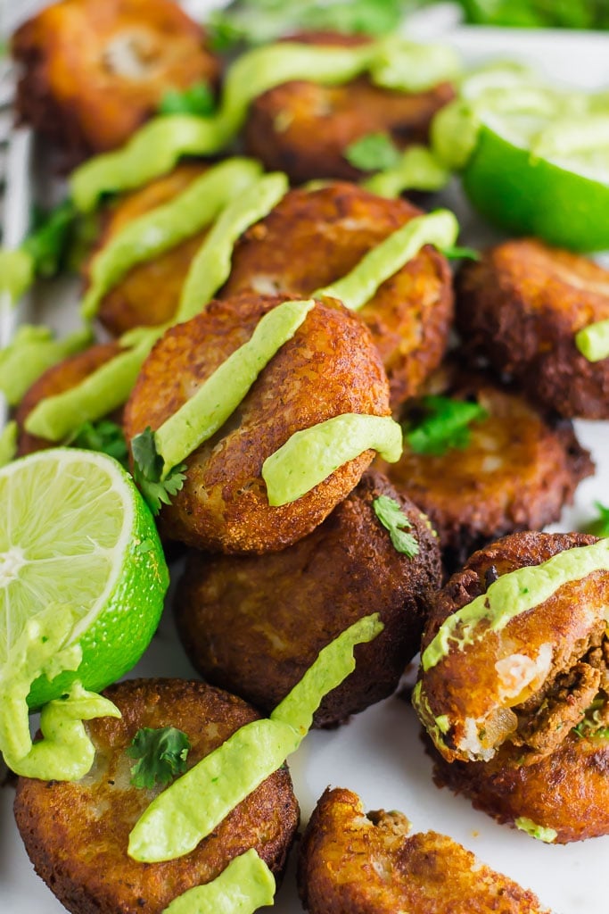 Fried mashed potato patties stuffed with taco seasoned ground beef and topped with avocado cream and fresh chopped cilantro on a white rectangular platter with fresh limes - fried potato and beef taco croquettes