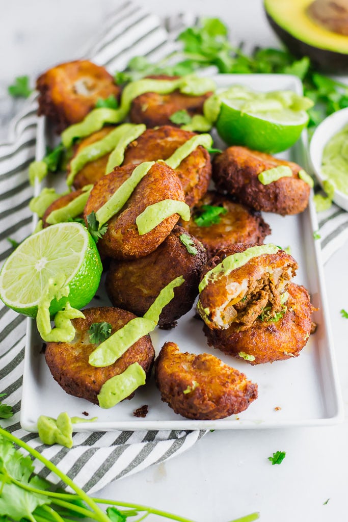 Fried mashed potato patties stuffed with taco seasoned ground beef and topped with avocado cream and fresh chopped cilantro on a white rectangular platter with fresh limes - fried potato and beef taco croquettes