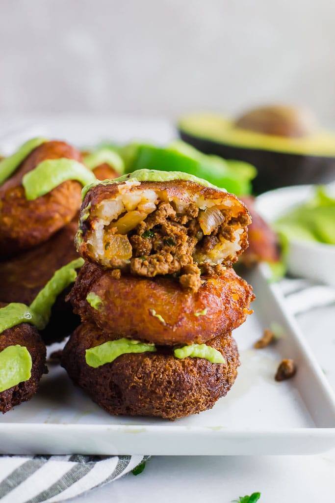 Fried mashed potato patties stuffed with taco seasoned ground beef and topped with avocado cream and fresh chopped cilantro on a white rectangular platter with fresh limes - fried potato and beef taco croquettes - gluten-free and dairy-free