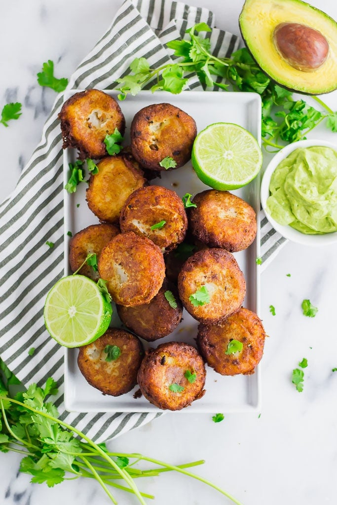 Fried mashed potato patties stuffed with taco seasoned ground beef and topped with avocado cream and fresh chopped cilantro on a white rectangular platter with fresh limes - fried potato and beef taco croquettes
