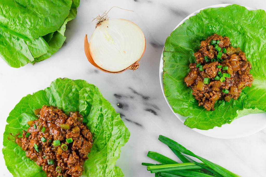 Sloppy Joe Lettuce Wraps - two gluten-free dairy-free sloppy joe lettuce wraps sitting on a marble table with fresh green onions and yellow onion and a pile of lettuce leaves - the ground beef is flavored with mustard, tomato sauce, tamari sauce, brown sugar, apple cider vinegar and topped with fresh chopped green onion