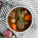A bowl of gluten-free beef stew with chunks of beef, carrots, potatoes, and fresh thyme on top, placed on a striped cloth with whole potatoes nearby.
