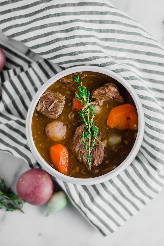 Whole30 Slow Cooker Beef Stew - a bowl of beef stew filled with red potatoes, carrots, and pearl onions in a dark beef broth in a white bowl topped with a sprig of fresh thyme sitting on top of a marble table with a white and blue striped napkin, a gold spoon, fresh herbs.