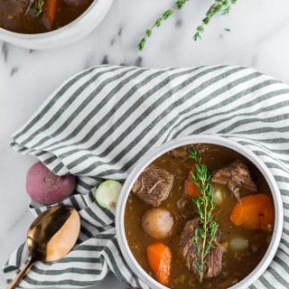 Whole30 Slow Cooker Beef Stew - a bowl of beef stew filled with red potatoes, carrots, and pearl onions in a dark beef broth in a white bowl topped with a sprig of fresh thyme sitting on top of a marble table with a white and blue striped napkin, a gold spoon, fresh herbs. Another bowl of beef stew in the top left corner.