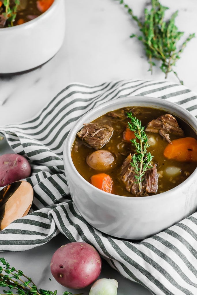 Whole30 Slow Cooker Beef Stew - a bowl of beef stew filled with red potatoes, carrots, and pearl onions in a dark beef broth in a white bowl topped with a sprig of fresh thyme sitting on top of a marble table with a white and blue striped napkin, a gold spoon, fresh herbs. Another bowl of beef stew in the top left corner.