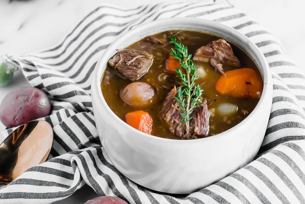 Whole30 Slow Cooker Beef Stew - a bowl of beef stew filled with red potatoes, carrots, and pearl onions in a dark beef broth in a white bowl topped with a sprig of fresh thyme sitting on top of a marble table with a white and blue striped napkin, a gold spoon, fresh herbs.