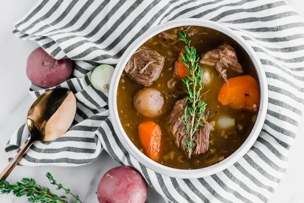 Whole30 Slow Cooker Beef Stew - a bowl of beef stew filled with red potatoes, carrots, and pearl onions in a dark beef broth in a white bowl topped with a sprig of fresh thyme sitting on top of a marble table with a white and blue striped napkin, a gold spoon, fresh herbs.