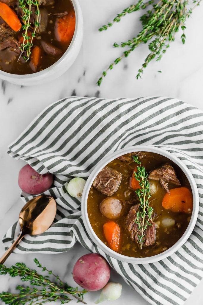 Whole30 Slow Cooker Beef Stew - a bowl of beef stew filled with red potatoes, carrots, and pearl onions in a dark beef broth in a white bowl topped with a sprig of fresh thyme sitting on top of a marble table with a white and blue striped napkin, a gold spoon, fresh herbs. Another bowl of beef stew in the top left corner.