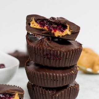 A stack of four chocolate peanut butter cups that are also filled with a red berry chia seed jam. The top peanut butter cup has a bite taken out of it. There is a spoonful of peanut butter in the back right corner and another peanut butter cup in the front left corner with a bite taken out of it. There is a white bowl sticking out on the side filled with jam.