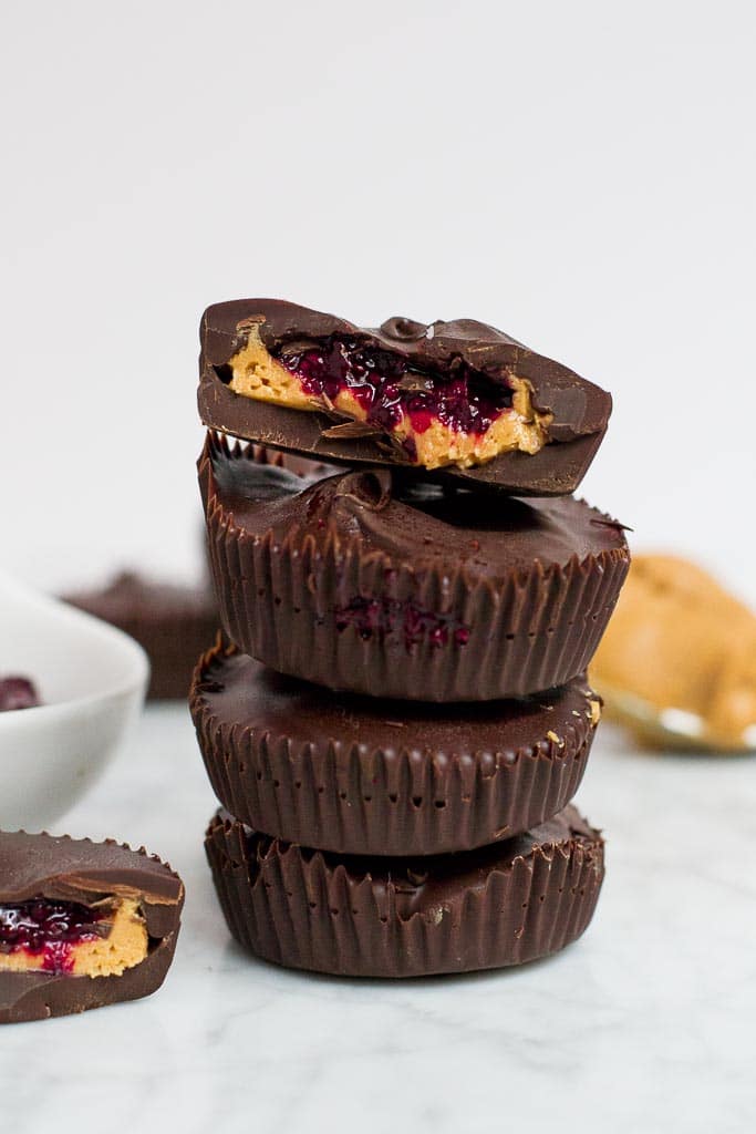 A stack of four chocolate peanut butter cups that are also filled with a red berry chia seed jam. The top peanut butter cup has a bite taken out of it. There is a spoonful of peanut butter in the back right corner and another peanut butter cup in the front left corner with a bite taken out of it. There is a white bowl sticking out on the side filled with jam. 