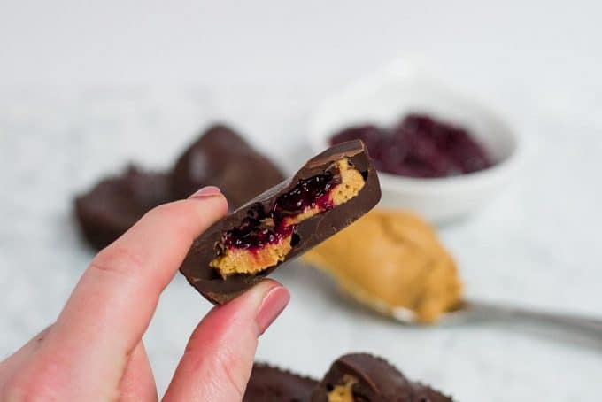 A hand holding half of a chocolate peanut butter cup that is also filled with a red berry chia jam. There is a spoonful of peanut butter and a bowl of red berry chia jam in the background. 