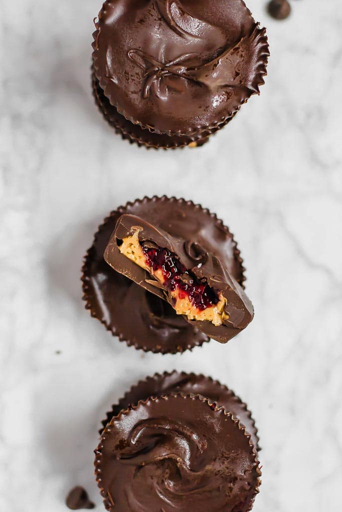 Three chocolate chia jam peanut butter cups in a vertical line sitting on a marble table. The middle chocolate cup is cut in half so you can see the peanut butter and red berry chia jam layers. 