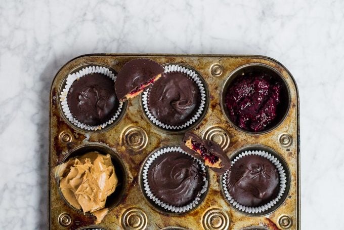 A muffin tin filled with chocolate peanut butter cups. There is one muffin well filled with a red berry chia jam. Another muffin well is filled with creamy peanut butter. There are half segments of chocolate peanut butter cups sitting on top of the muffin tin so you can see the layers inside. 
