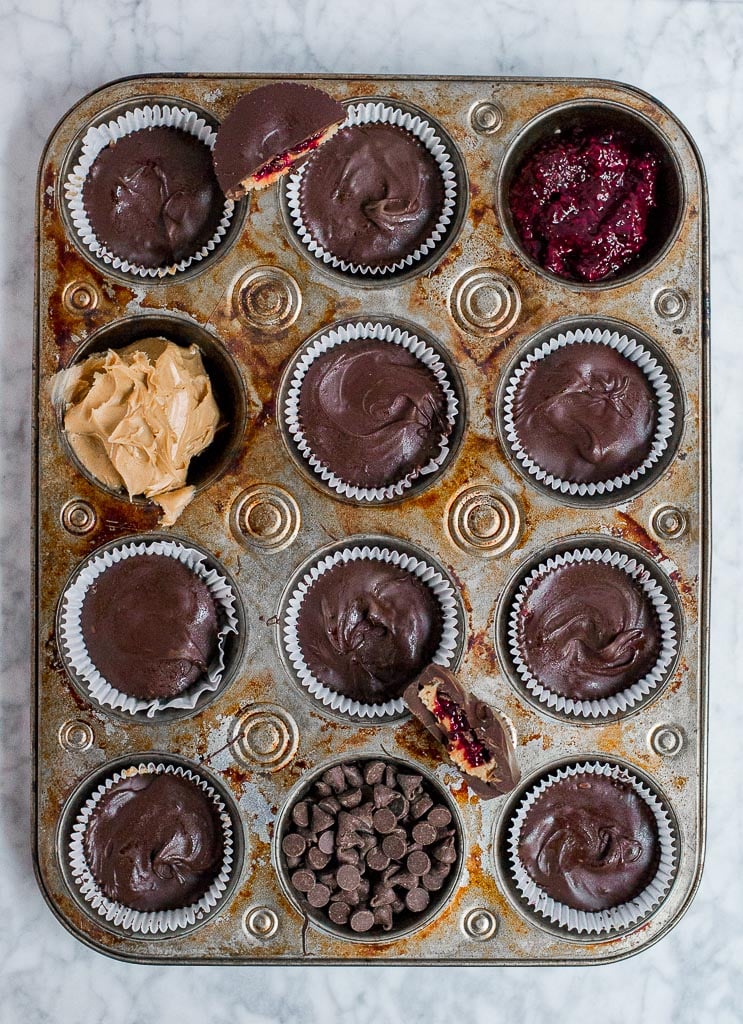 A muffin tin filled with chocolate peanut butter cups. There is one muffin well filled with a red berry chia jam. Another muffin well is filled with creamy peanut butter. There are half segments of chocolate peanut butter cups sitting on top of the muffin tin so you can see the layers inside.