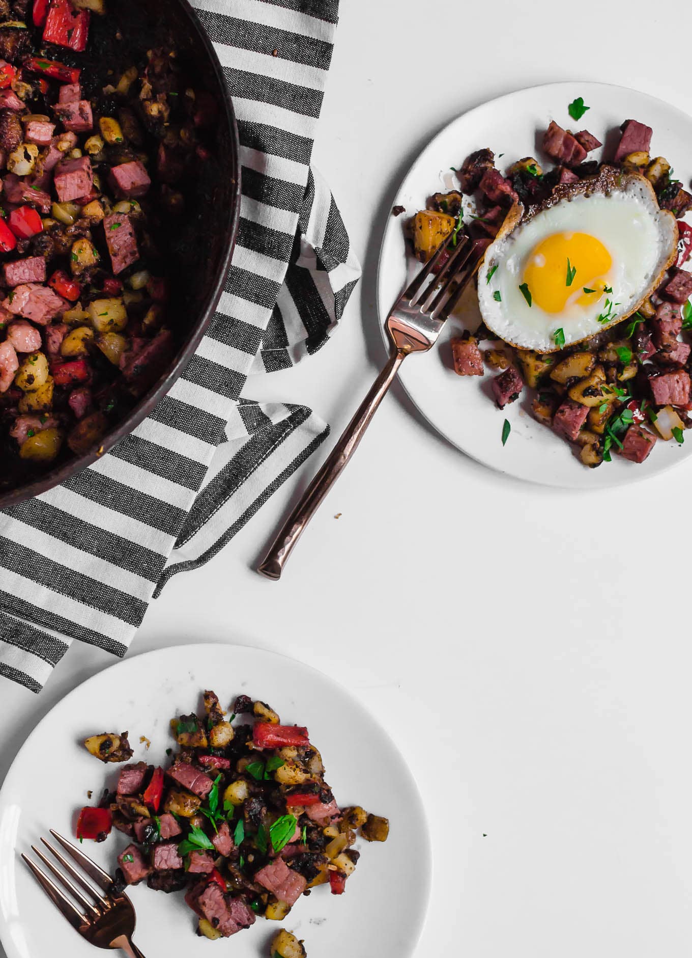 An aerial view of two white plates holding gluten-free corned beef hash, one with an egg on top. Two forks ready to dig in to each plate of food and a large skillet holding the remaining leftover corned beef hash. Everything is sitting on a white table with a grey and white napkin. 