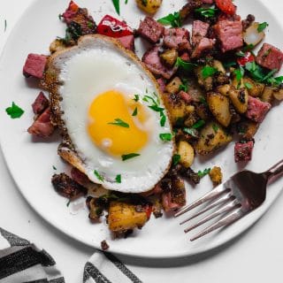 An aerial view of a white plate of gluten-free leftover corned beef hash topped with a sunny side up egg sitting on a white table, with a fork and a gray and white napkin underneath it.