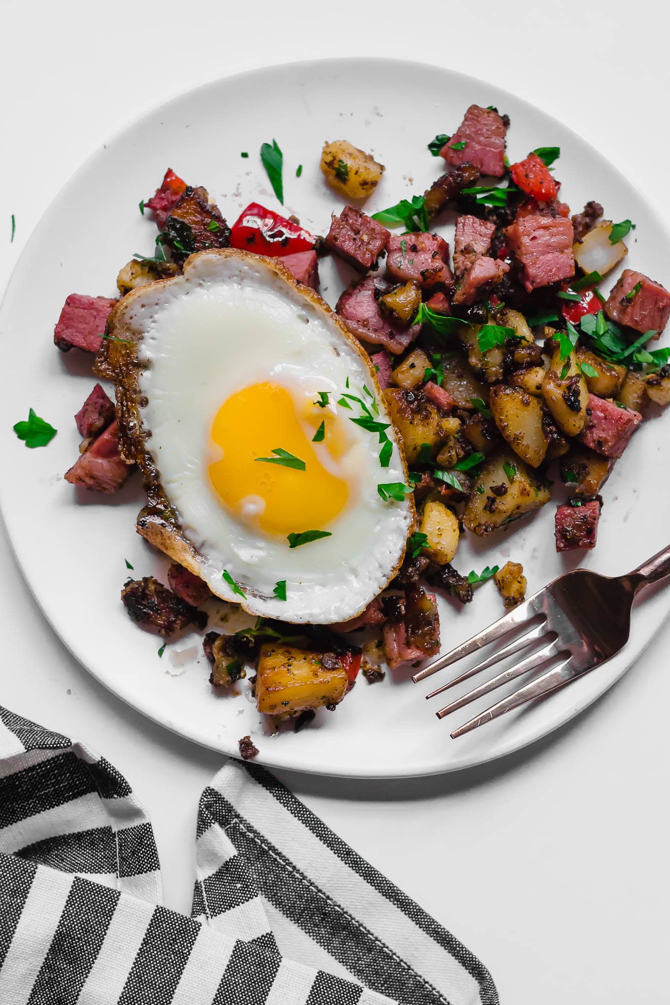 An aerial view of a white plate of gluten-free leftover corned beef hash topped with a sunny side up egg sitting on a white table, with a fork and a gray and white napkin underneath it. 
