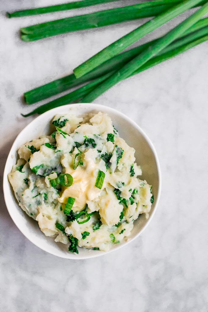 A bowl of dairy-free kale colcannon - mashed potatoes in a white bowl mixed with dark green kale and green onions, topped with a pat of dairy-free butter on a marble table with fresh green onions laying on the table above the bowl