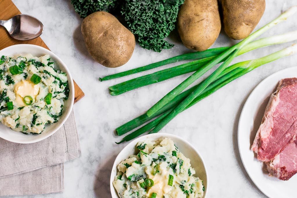 two bowls of dairy-free kale colcannon on a marble table with russet potatoes, green kale, corned beef and green onions