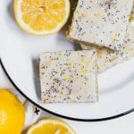 An aerieal view of a white and black plate filled with lemon poppy seed cookie bars topped with lemon glaze and poppy seeds and fresh lemons.