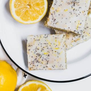 An aerieal view of a white and black plate filled with lemon poppy seed cookie bars topped with lemon glaze and poppy seeds and fresh lemons.