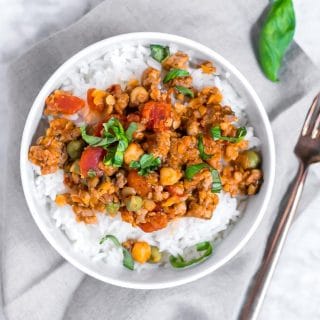 Italian sausage, lentil and chickpeas in a tomato sauce topped with basil in a bowl of white rice sitting on a gray napkin on a marble table with a gold fork on the side of it