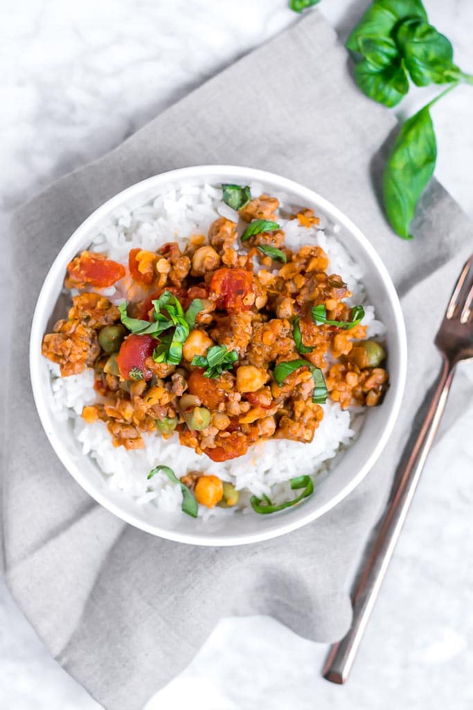 one pot garbanzo lentil and italian sausage - Italian sausage, lentil and chickpeas in a tomato sauce topped with basil in a bowl of white rice sitting on a gray napkin on a marble table with a gold fork on the side of it