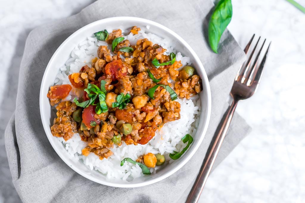 one pot garbanzo lentil and italian sausage - Italian sausage, lentil and chickpeas in a tomato sauce topped with basil in a bowl of white rice sitting on a gray napkin on a marble table with a gold fork on the side of it