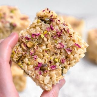 A hand holding a gluten-free marshmallow treat made from rice cereal and topped with pistachios and rose petals against a white background with more marshmallow treats in the distance sitting on a marble table