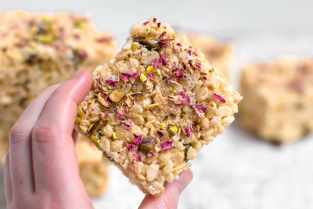 A hand holding a gluten-free marshmallow treat made from rice cereal and topped with pistachios and rose petals against a white background with more marshmallow treats in the distance sitting on a marble table