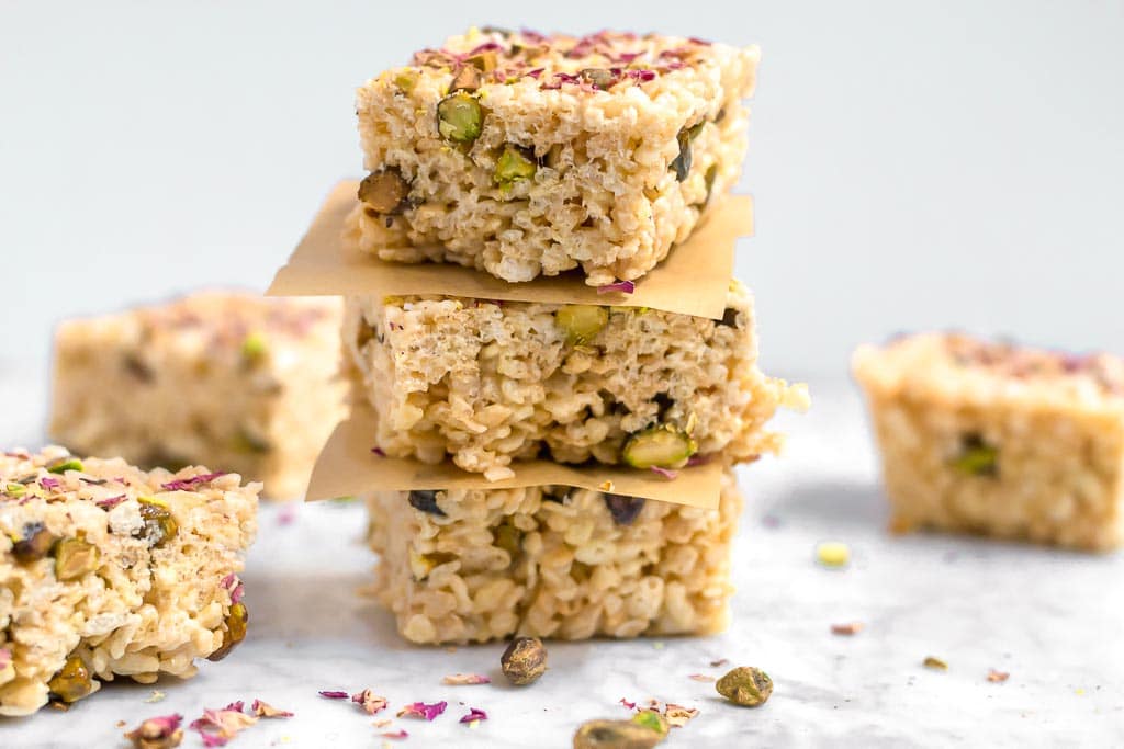 A stack of three gluten-free marshmallow treats made from rice cereal and topped with pistachios and rose petals on a marble table. Parchment paper squares separates each marshmallow treat. there are pistachios and rose petals scattered on the table.
