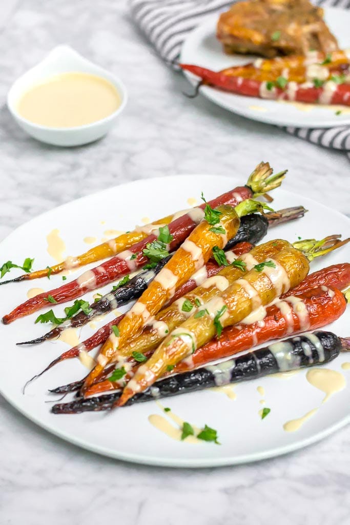 Multi-color roasted rainbow carrots on a white plate drizzled with a light and creamy lemon tahini sauce. A small bowl of sauce in the upper left corner. Roasted Rainbow Carrots with Lemon Tahini Sauce - Gluten-free, Dairy-free, Vegan - A Dash of Megnut