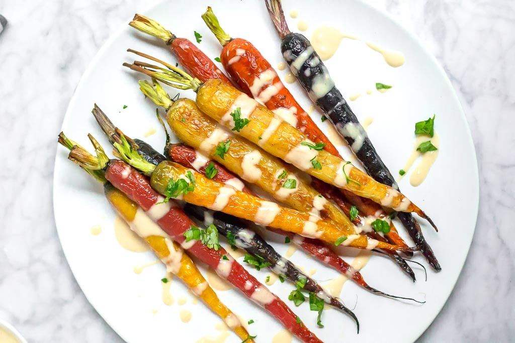 Multi-color roasted rainbow carrots on a white plate drizzled with a light and creamy lemon tahini sauce. A small bowl of sauce in the upper left corner. Roasted Rainbow Carrots with Lemon Tahini Sauce - Gluten-free, Dairy-free, Vegan - A Dash of Megnut