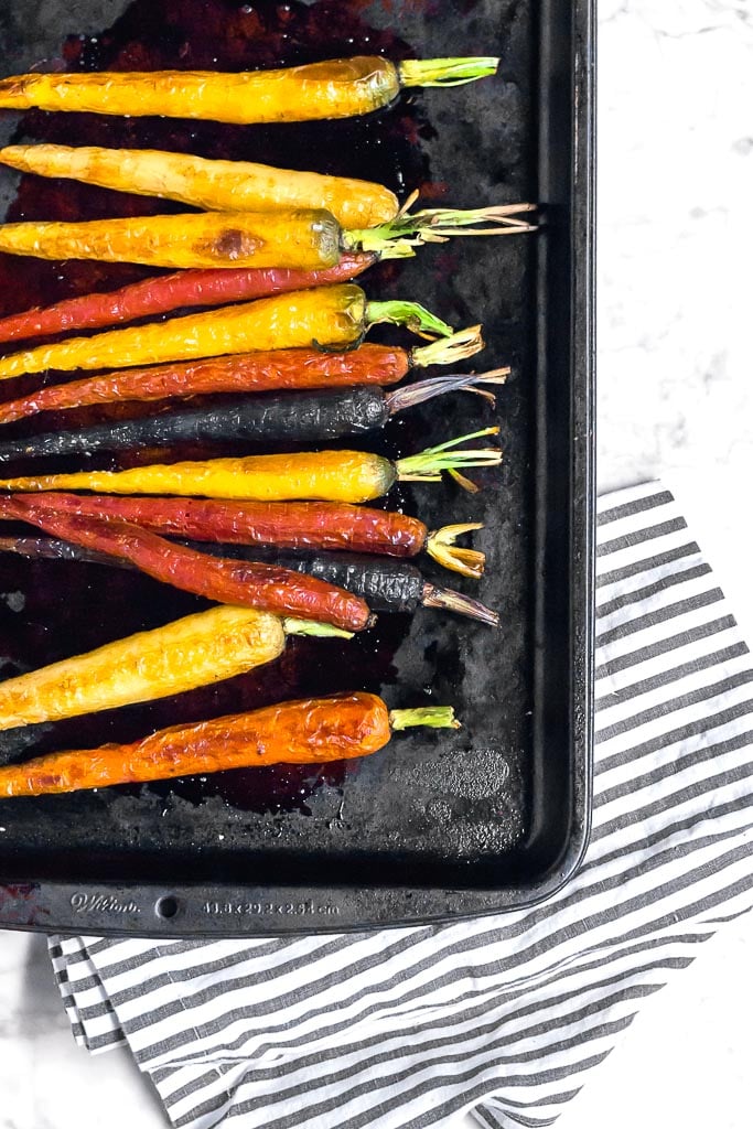 A baking sheet with roasted multi-color rainbow carrots on it sitting on a marble table with a white and gray striped towel. 