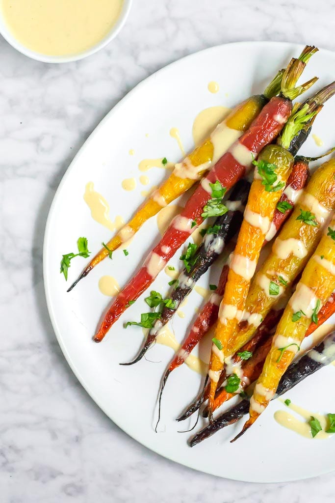 Multi-color roasted rainbow carrots on a white plate drizzled with a light and creamy lemon tahini sauce. A small bowl of sauce in the upper left corner. Roasted Rainbow Carrots with Lemon Tahini Sauce - Gluten-free, Dairy-free, Vegan - A Dash of Megnut