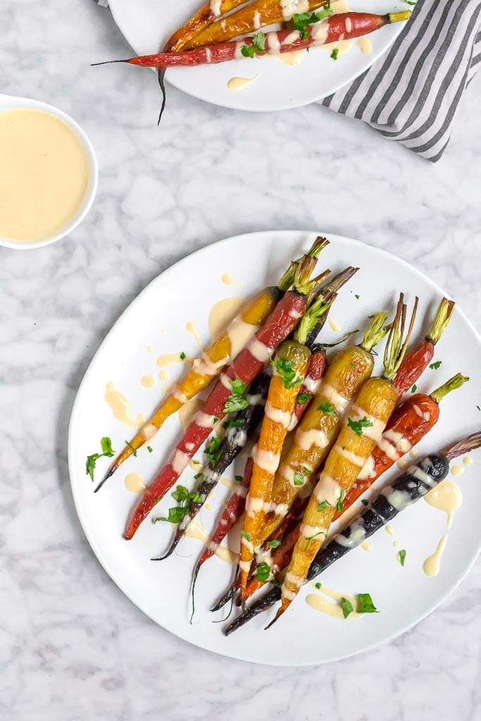 Multi-color roasted rainbow carrots on a white plate drizzled with a light and creamy lemon tahini sauce. A small bowl of sauce in the upper left corner. Roasted Rainbow Carrots with Lemon Tahini Sauce - Gluten-free, Dairy-free, Vegan - A Dash of Megnut