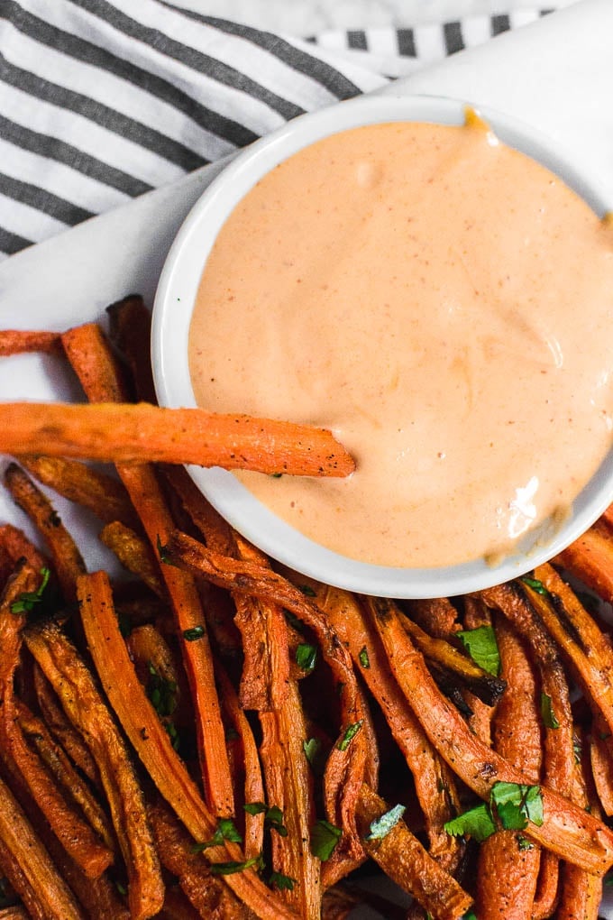 An aerial view of a plate of baked carrot fries with a fry dipped into sriracha mayo. 