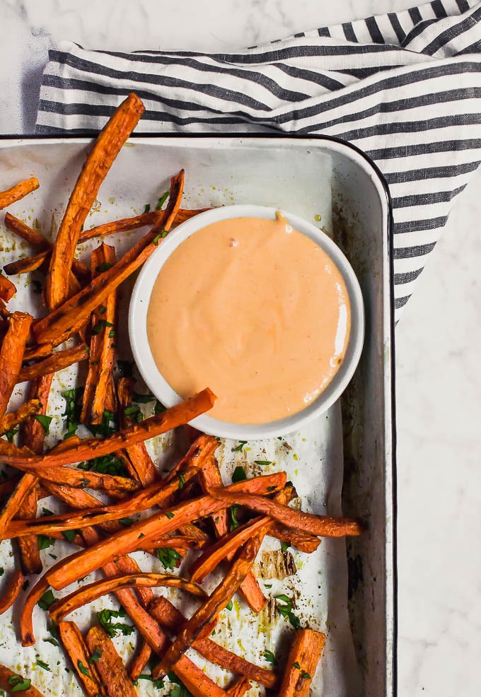 Aerial view of baked carrot fries on a baking sheet topped with parsley and a bowl with sriracha mayo.