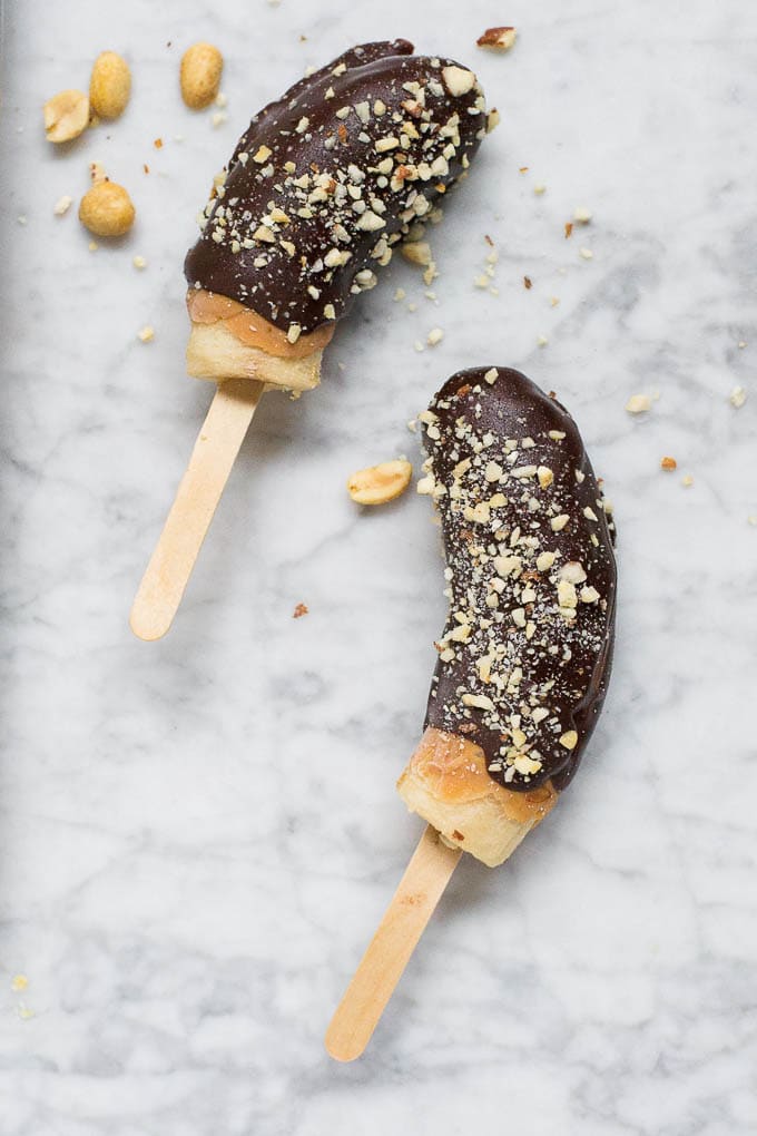 An aerial view of two frozen bananas covered with peanut butter, chocolate and chopped roasted salted peanuts on a marble table. 